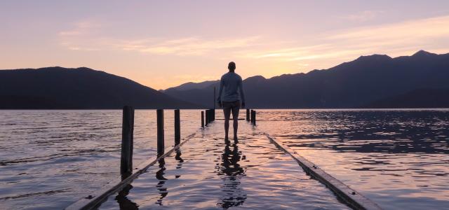 Silhouette of a person from behind standing at the end of a dock looking out at a lake. The sun is rising behind the mountains.